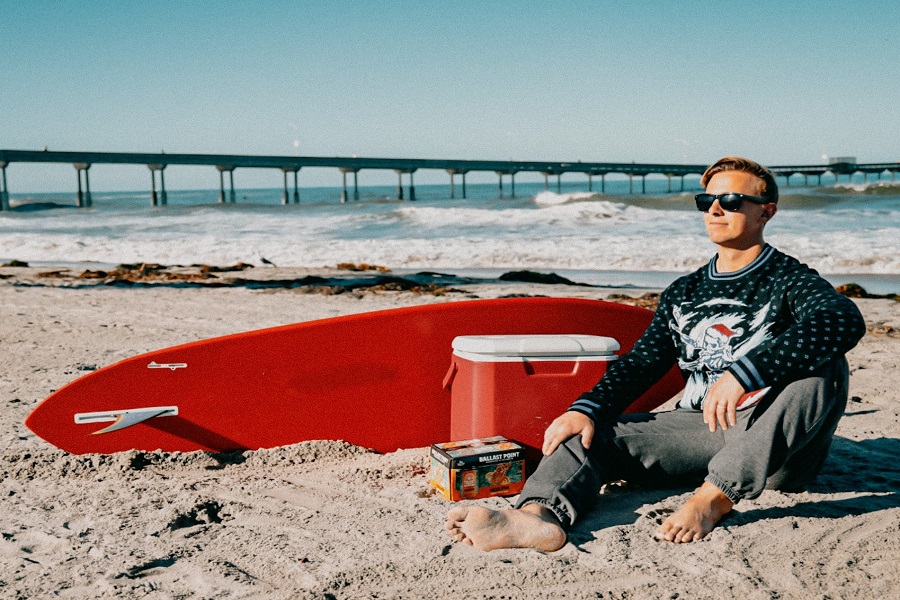 man with a surfboard and cooler sitting on a beach wearing an ugly sweater
